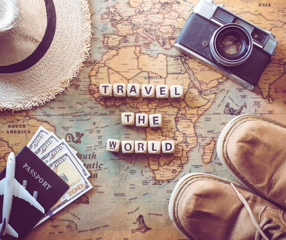 A vintage travel-themed flat lay composition featuring a world map with the words 'TRAVEL THE WORLD' spelled out using wooden letter blocks. Surrounding the map are travel essentials including a straw hat, an old-fashioned camera, a passport with U.S. dollar bills, a small toy airplane, and a pair of worn brown boots, symbolizing adventure and exploration.
